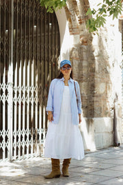 Person wearing a white dress, light blue cardigan, and brown boots standing under a tree.