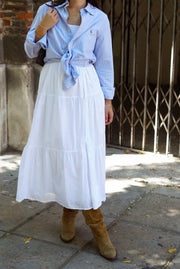 Person wearing a blue shirt tied around the waist with a white skirt and brown boots, standing on a stone pavement.
