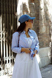 Woman wearing a blue cap and light shirt with a white skirt standing against a stone wall.