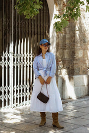 Woman in a light blue shirt, white skirt, and brown boots standing in front of a stone wall.