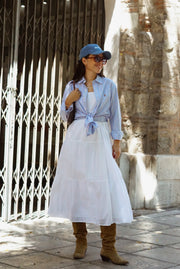 Woman in a white dress and blue shirt with rolled-up sleeves standing in front of a stone wall.