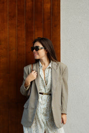 Woman in a beige blazer over a floral dress standing against a wooden and white wall.