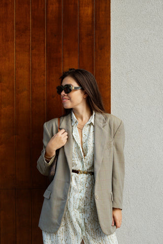 Woman in a beige blazer over a floral dress standing against a wooden and white wall.